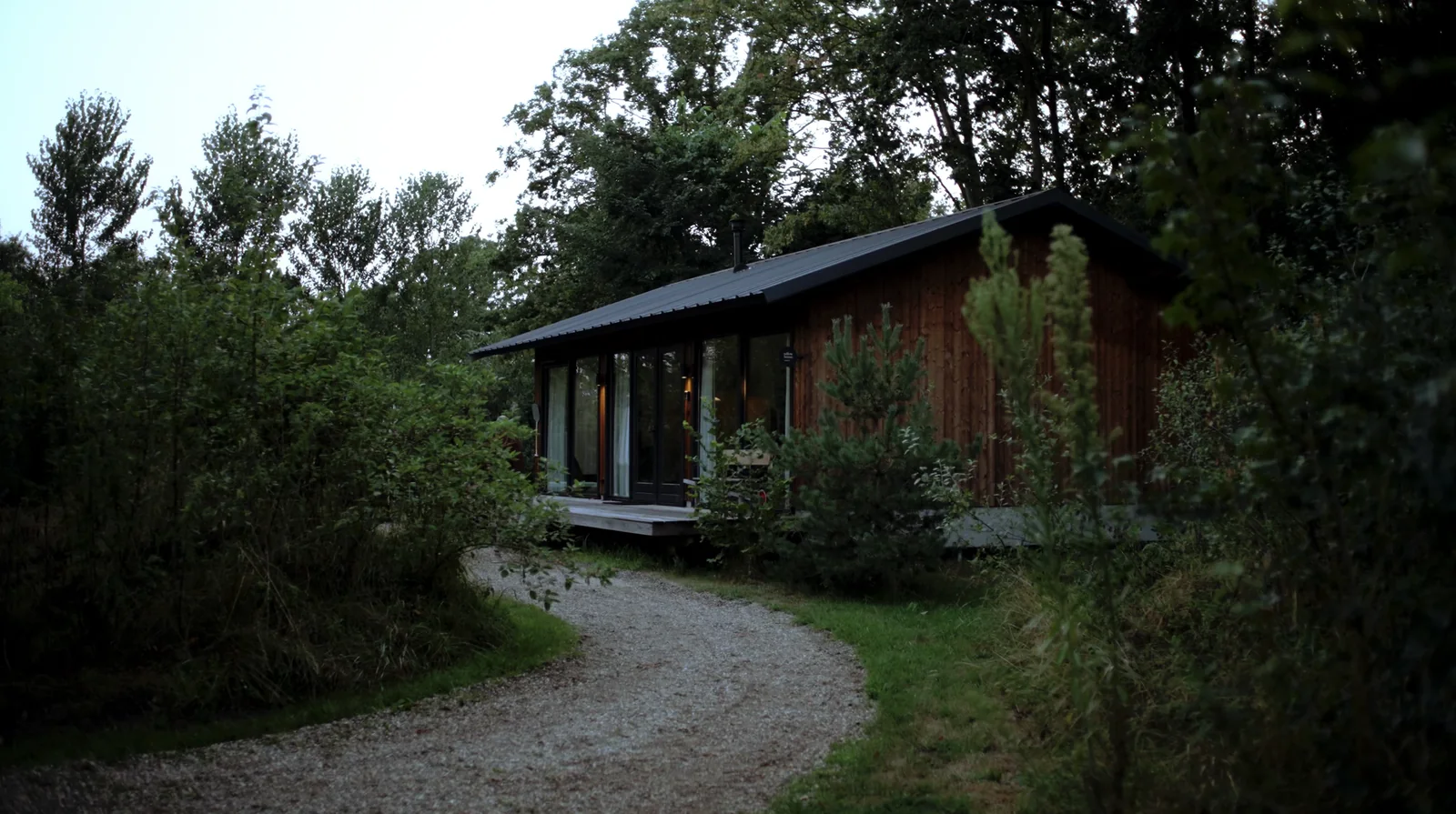 A wooden house among trees at dusk, a gravel path leading to it.