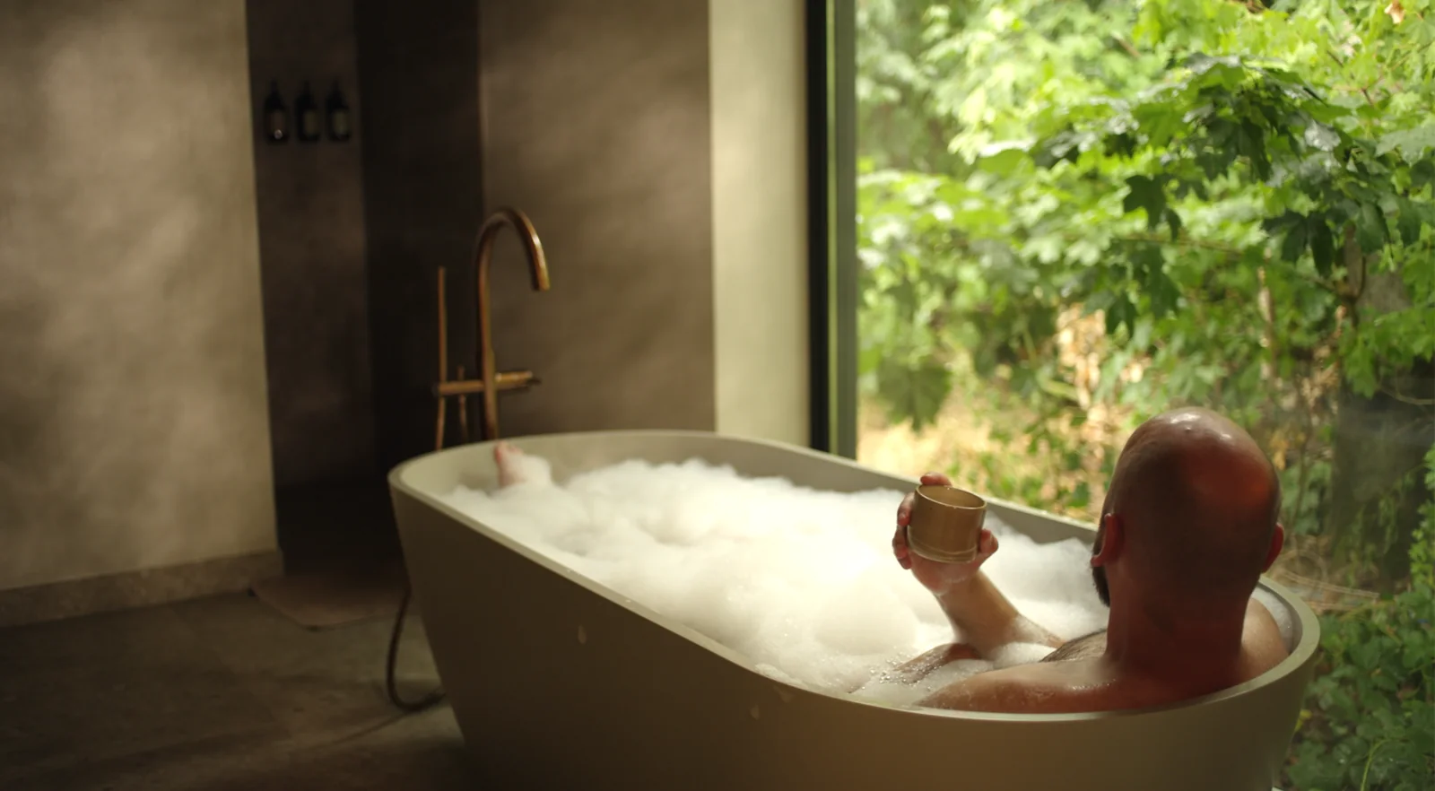 A man resting in a bath with a cup of tea, trees outside the window.